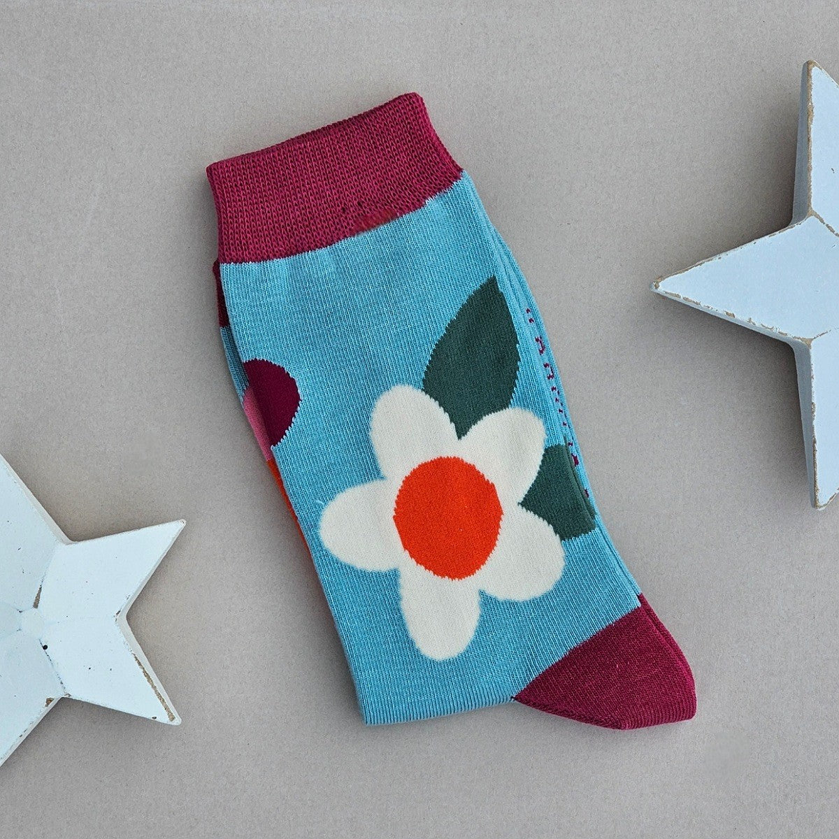 A single sock laid flat on a light grey surface, decorated with a large white/orange and deep red flowers and dark green leaves on an duck egg base, with deep red cuff, heel and toes. Two white star-shaped props are partially visible at the edges.