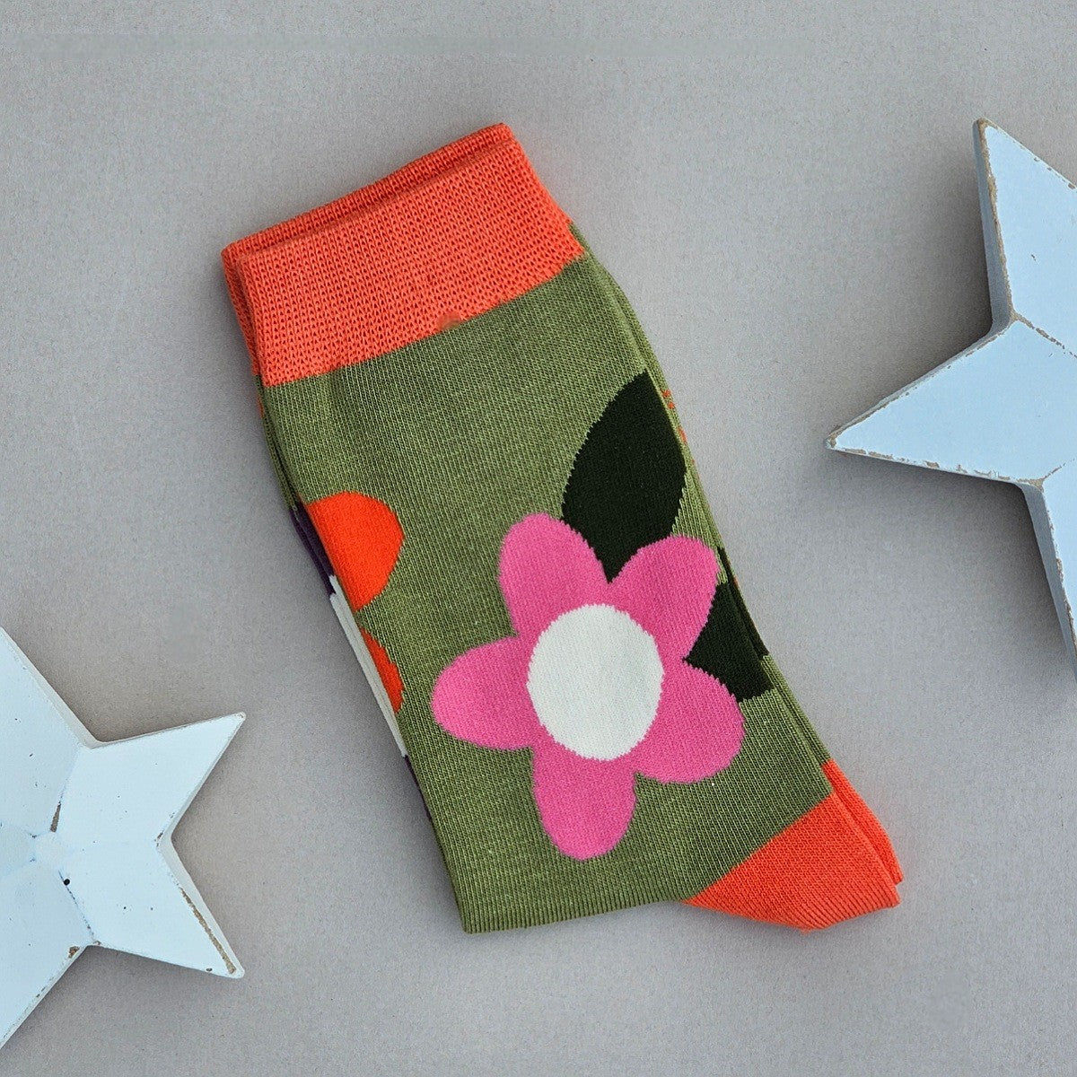 A single sock laid flat on a light grey surface, decorated with a large pink and orange flowers and dark green leaves on an olive green base, with bright orange cuff, heel and toes. Two white star-shaped props are partially visible at the edges.