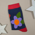 A single sock laid flat on a beige background with white star-shaped props. The sock is dark navy blue with a large purple flower and orange center on the front, plus red cuffs, heel and green leaves.