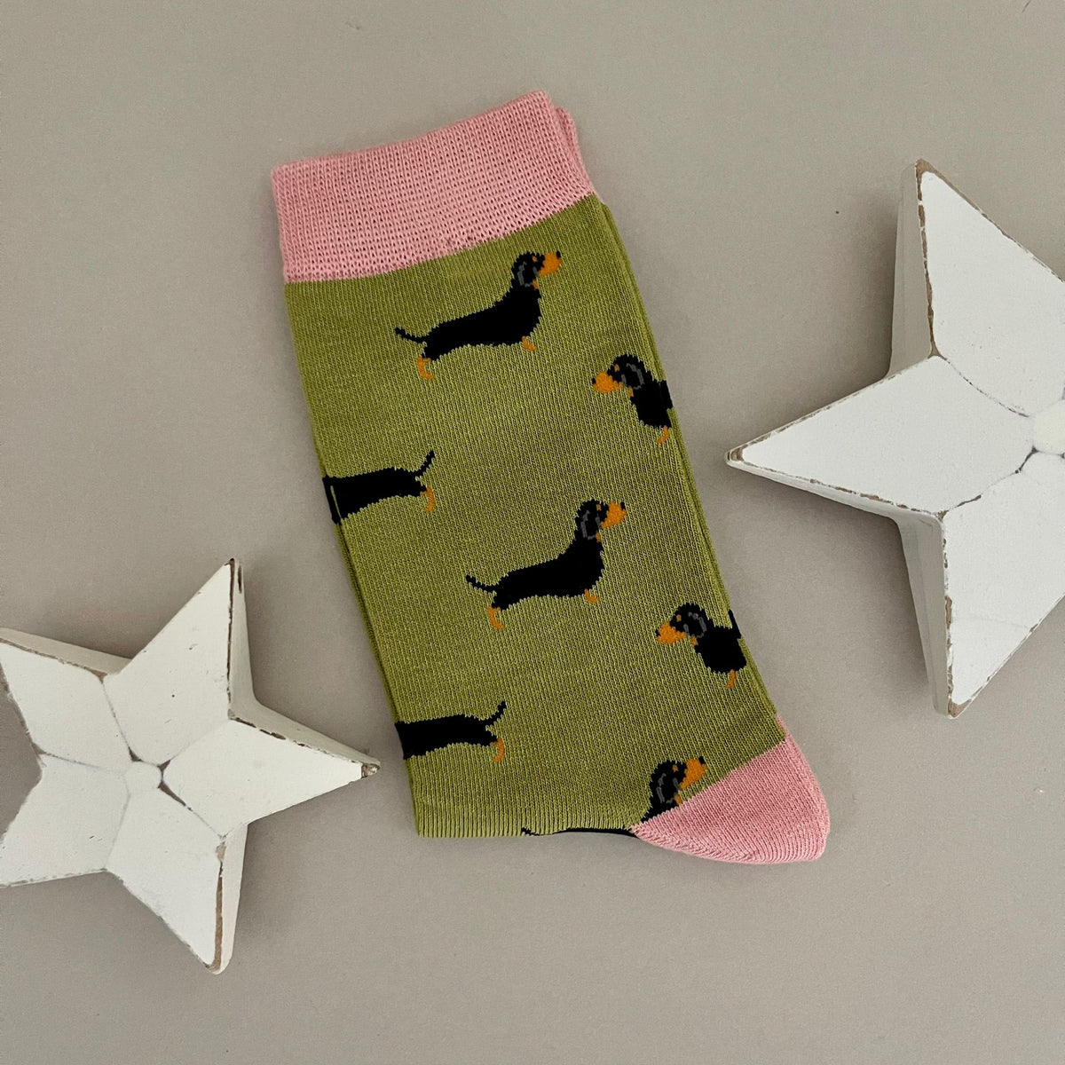 A single moss green sock with a repeating pattern of black little sausage dogs with orange feet and noses.  Featuring pink cuff, heel and toes, displayed on a neutral beige background with white star-shaped props