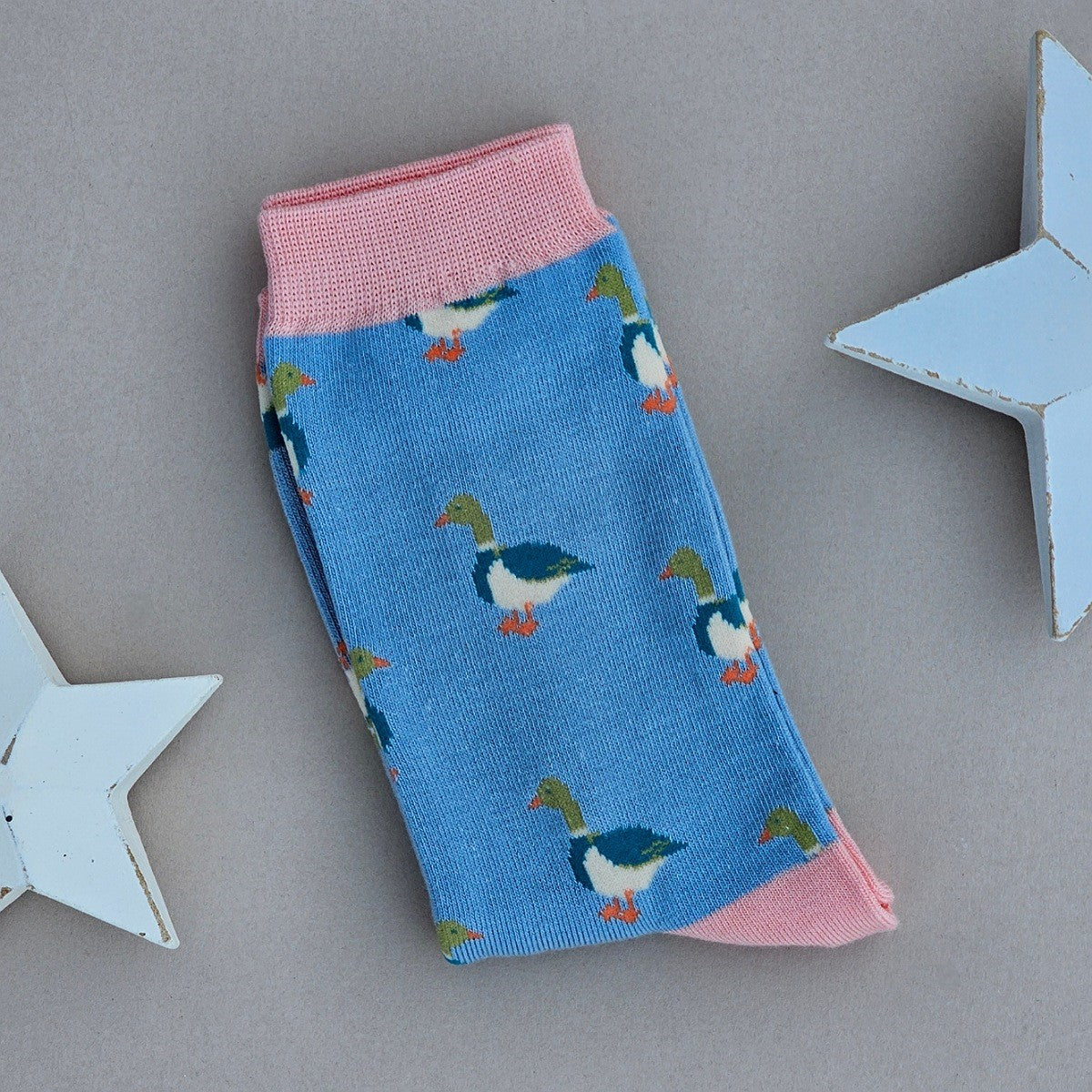 A single blue sock with various mallard duck silhouettes and pink yellow cuff, heel, and toe, laid flat on a light beige background with two white star decorations.