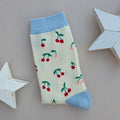 A single folded sock with a cream body, light blue cuffs, heel and toe with multiple red and pink cherry patterns, displayed on a neutral grey background with decorative white stars.
