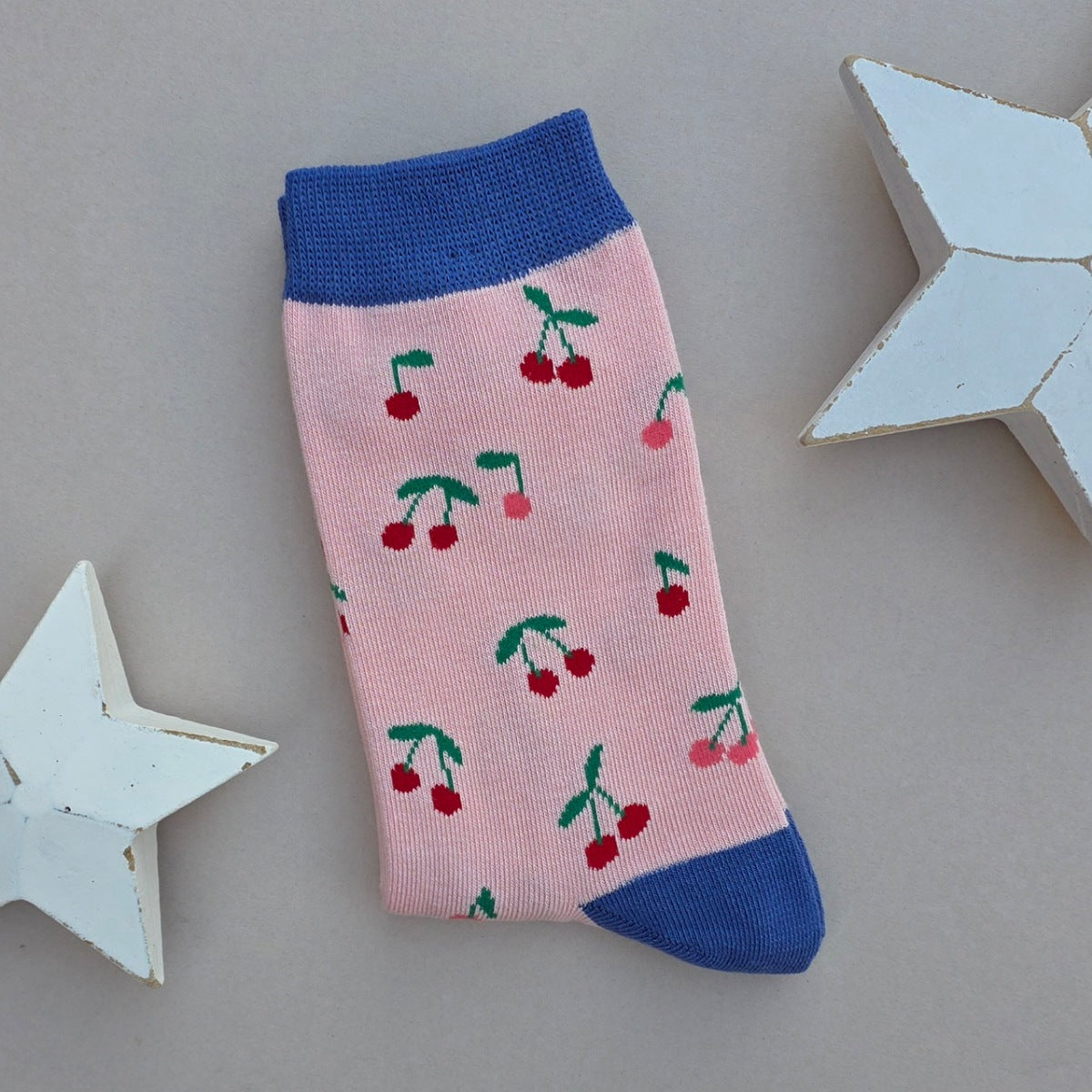 A single folded sock with a pink body, blue cuffs, heel and toe with multiple red, green and pink cherry patterns, displayed on a neutral grey background with decorative white stars.