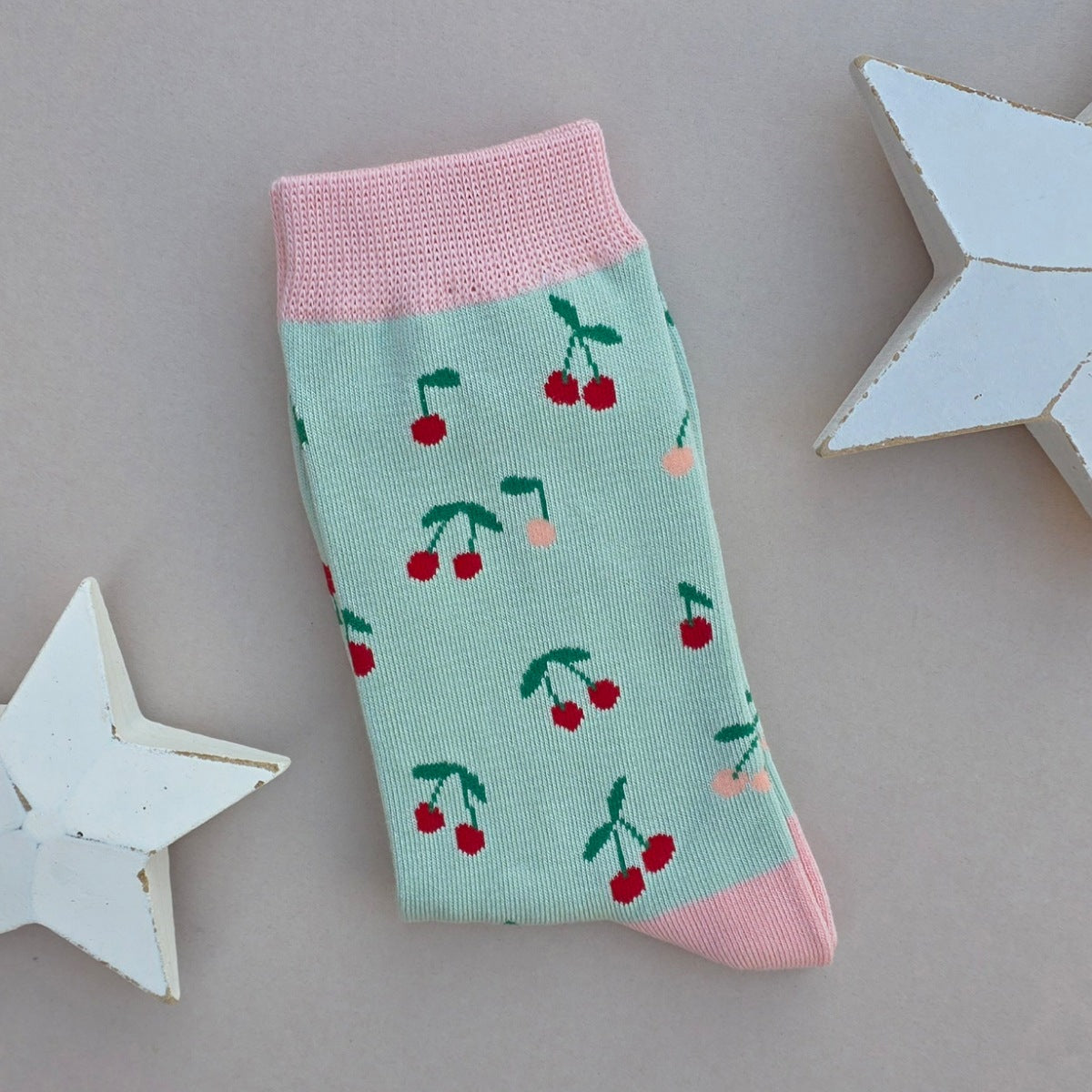 A single folded sock with a mint green body, pink cuffs, heel and toe with multiple red and pink cherry patterns, displayed on a neutral grey background with decorative white stars.