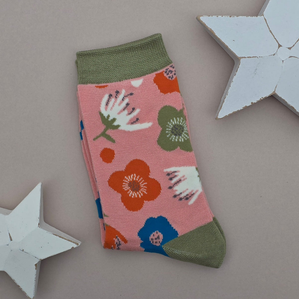 A single folded sock with a pink base and a colourful floral pattern, featuring an olive green cuff, heel and toe, displayed on a neutral beige background with decorative white stars.