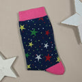 A single navy blue sock with a colourful star pattern and bright pink cuffs and heel, laid flat on a beige background with white star-shaped props.