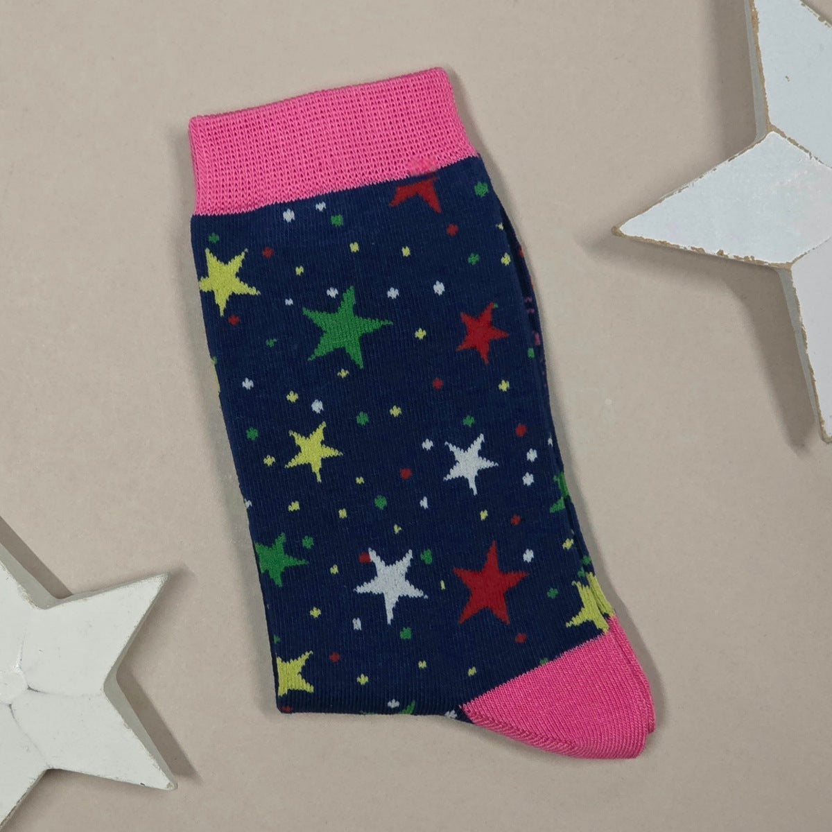 A single navy blue sock with a colourful star pattern and bright pink cuffs and heel, laid flat on a beige background with white star-shaped props.