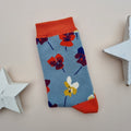A single blue knit sock with a colourful pattern of viola flowers, orange cuffs, heel and toes.  Displayed on a light beige background with decorative white star props.
