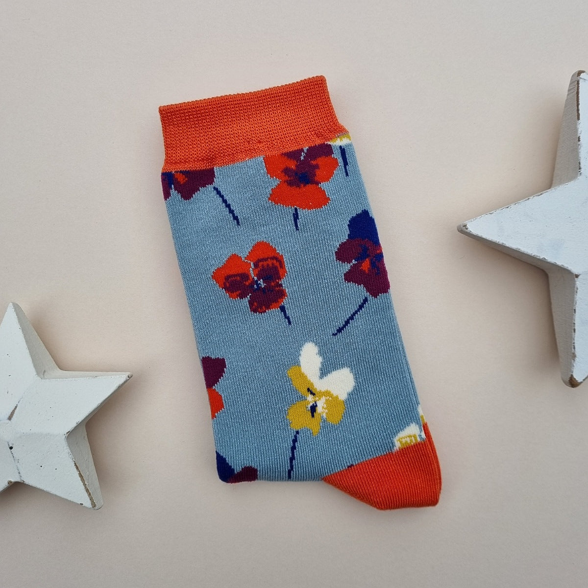 A single blue knit sock with a colourful pattern of viola flowers, orange cuffs, heel and toes.  Displayed on a light beige background with decorative white star props.