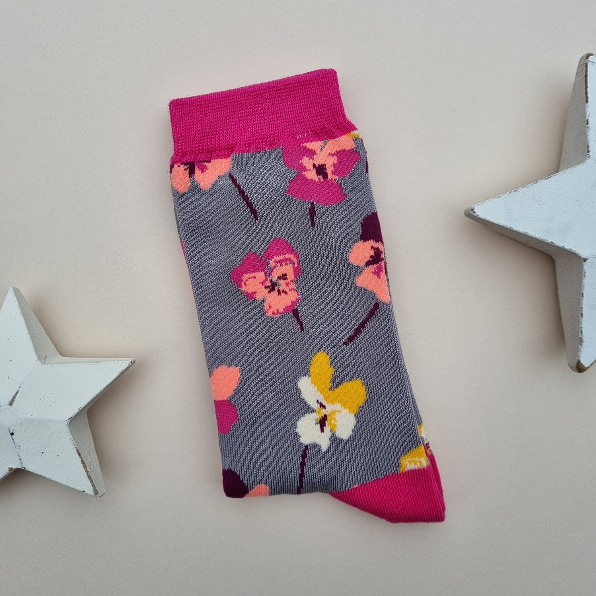 A single grey knit sock with a colourful pattern of viola flowers, bright pink cuffs, heel and toes.  Displayed on a light beige background with decorative white star props.