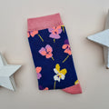 A single navy blue knit sock with a colourful pattern of viola flowers, pink cuffs, heel and toes.  Displayed on a light beige background with decorative white star props.