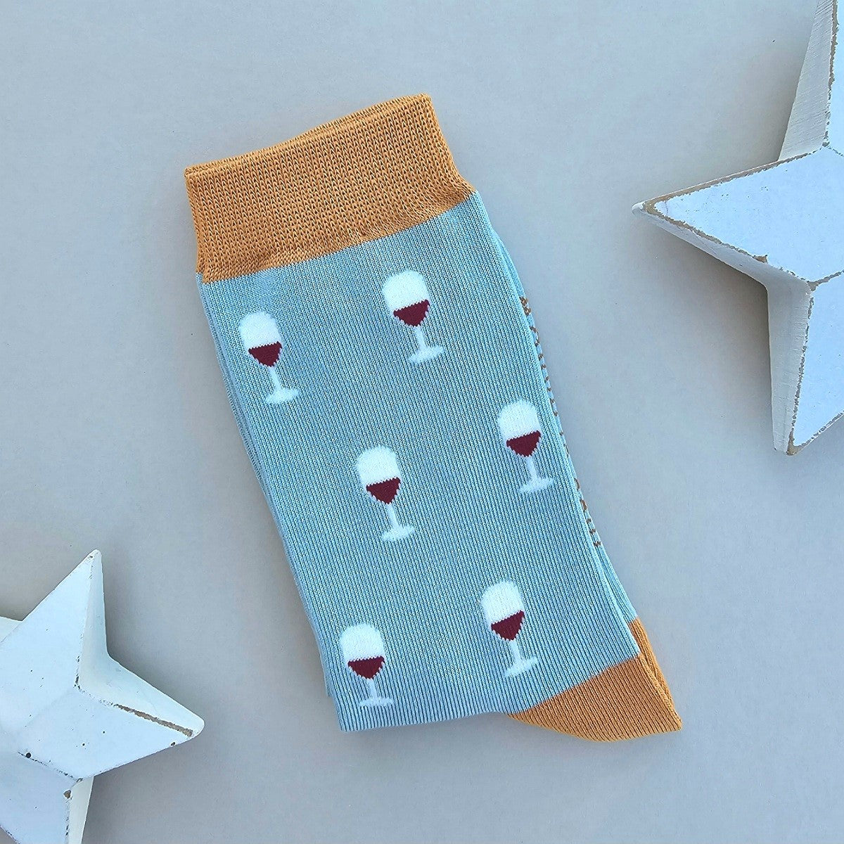 A single duck egg blue sock with an all-over pattern of white wine glasses filled with red wine.  Mustard ribbed cuffs and heels, styled on a light grey surface with white star props.
