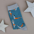 A single duck egg blue knit sock with light blue cuffs, toe and heel, featuring a pattern of women in orange and blue outfits performing yoga poses, displayed on a neutral grey background with white star-shaped props.