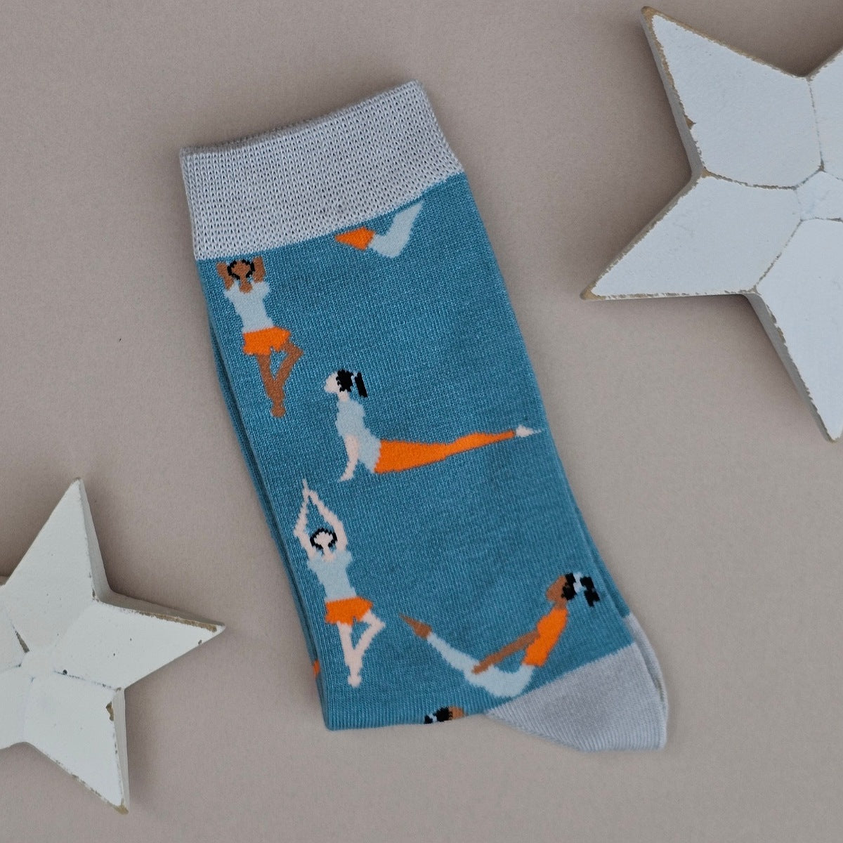 A single duck egg blue knit sock with light blue cuffs, toe and heel, featuring a pattern of women in orange and blue outfits performing yoga poses, displayed on a neutral grey background with white star-shaped props.