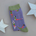 A single purple knit sock with green cuffs, toe and heel, featuring a pattern of women in orange and green outfits performing yoga poses, displayed on a neutral grey background with white star-shaped props.