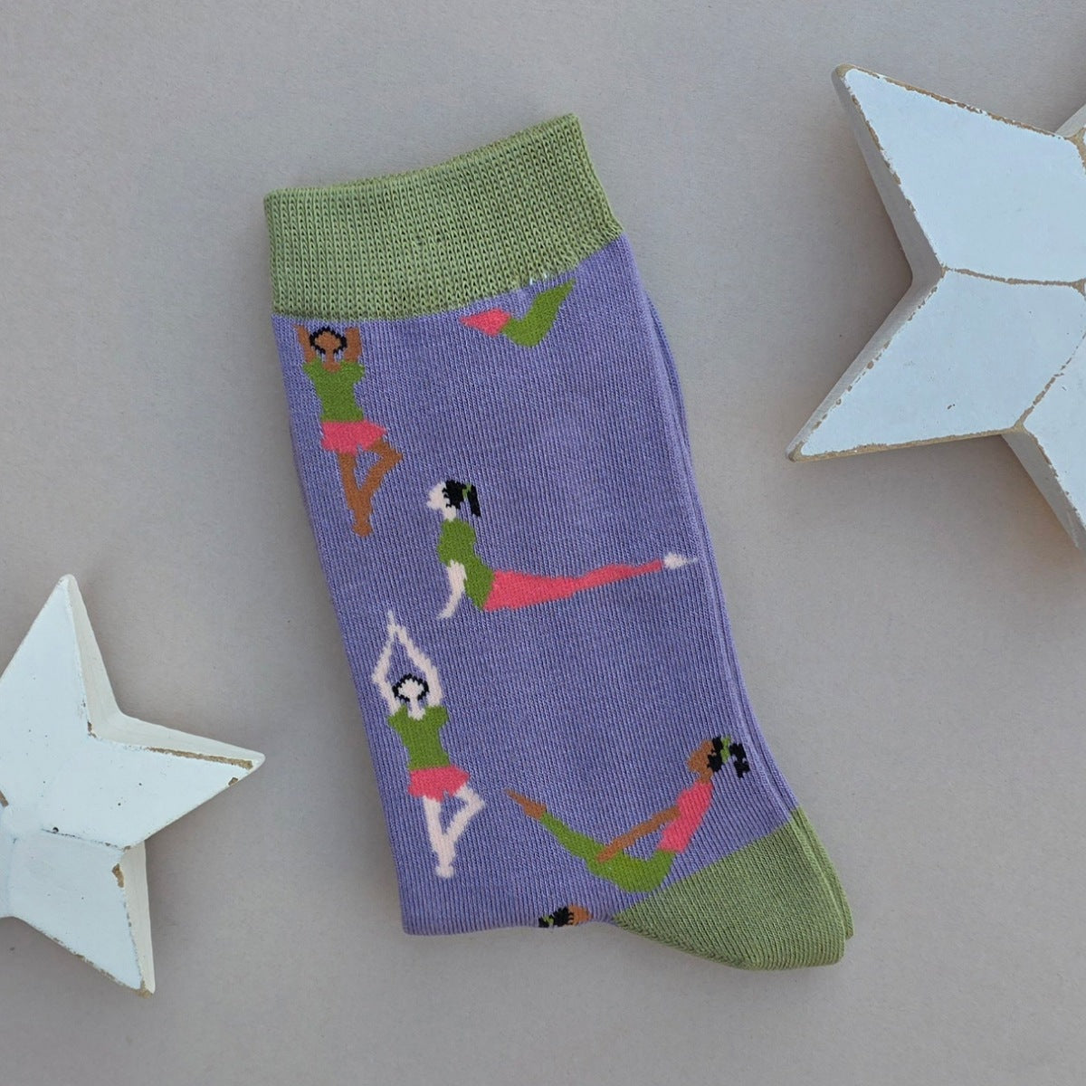 A single purple knit sock with green cuffs, toe and heel, featuring a pattern of women in orange and green outfits performing yoga poses, displayed on a neutral grey background with white star-shaped props.