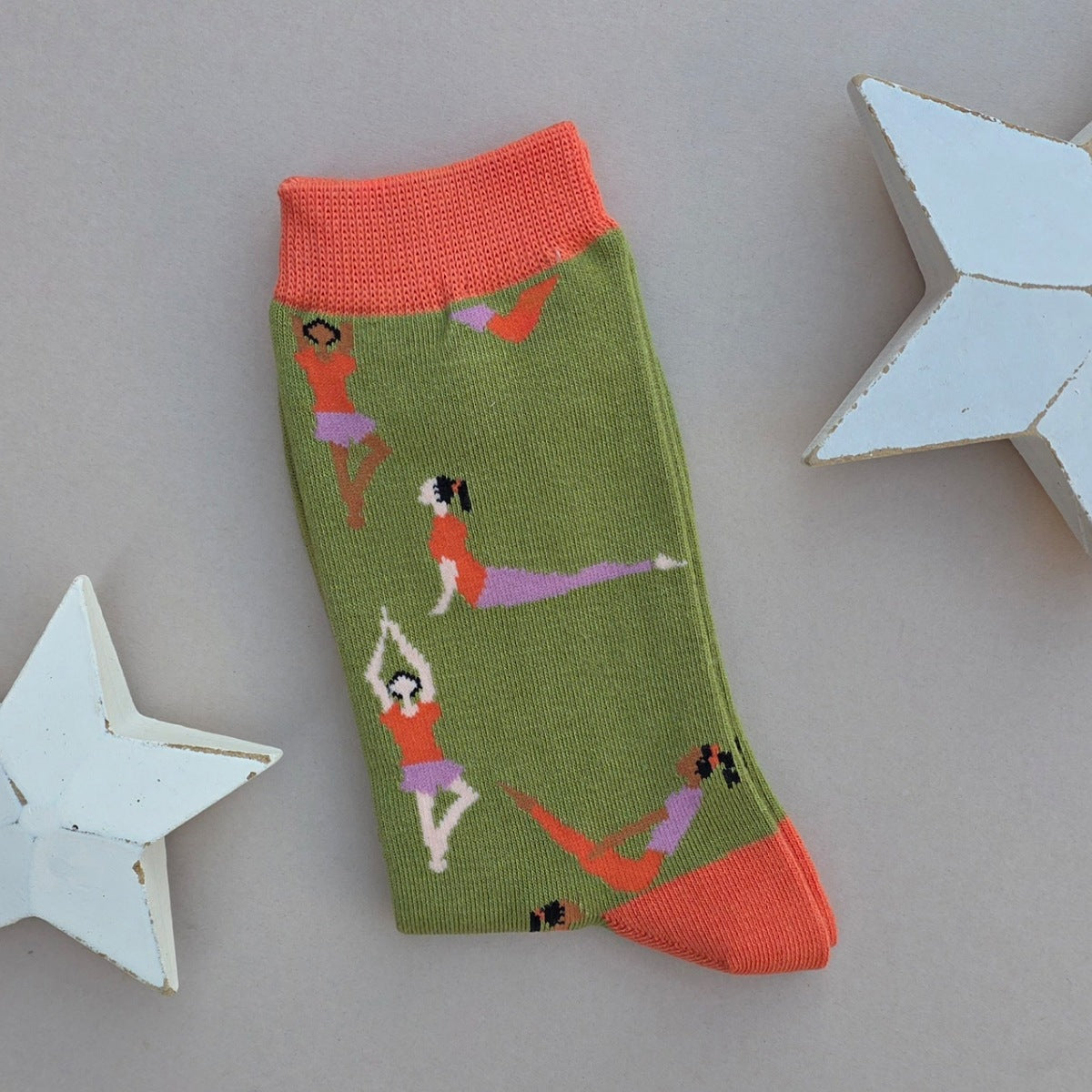 A single green knit sock with orange cuffs, toe and heel, featuring a pattern of women in orange and purple outfits performing yoga poses, displayed on a neutral grey background with white star-shaped props.