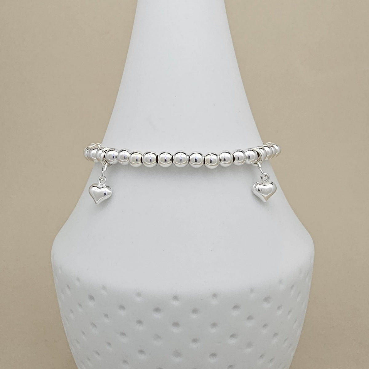 A silver beaded bracelet with two dangling heart charms, displayed on a white ceramic jewelry cone against a beige background.