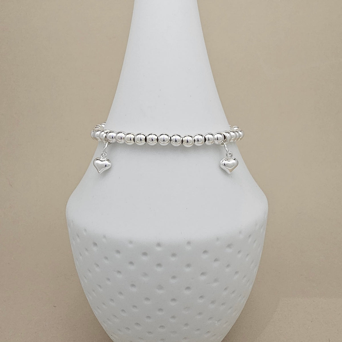 A silver beaded bracelet with two dangling heart charms, displayed on a white textured cone stand against a warm beige background.