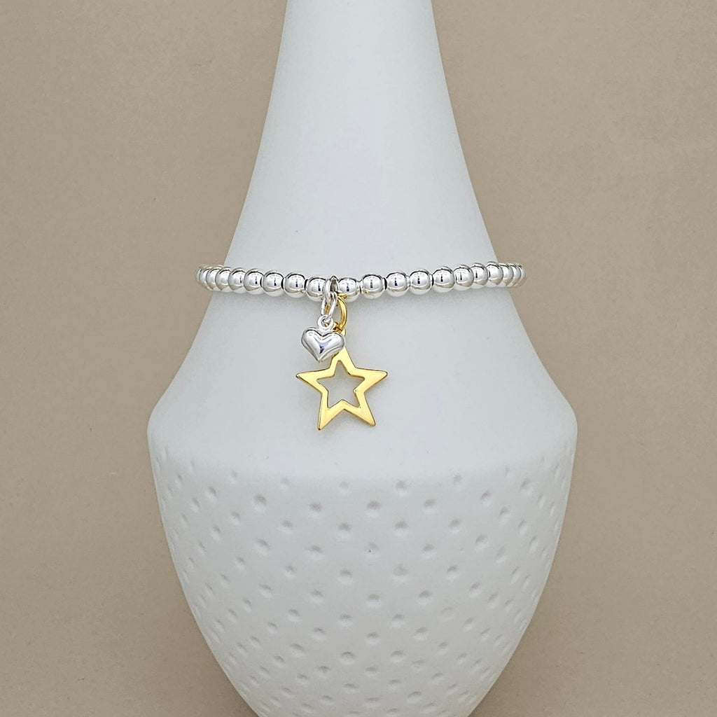 A silver beaded bracelet with a dangling gold star and silver heart charm, displayed on a white textured cone stand against a beige background.