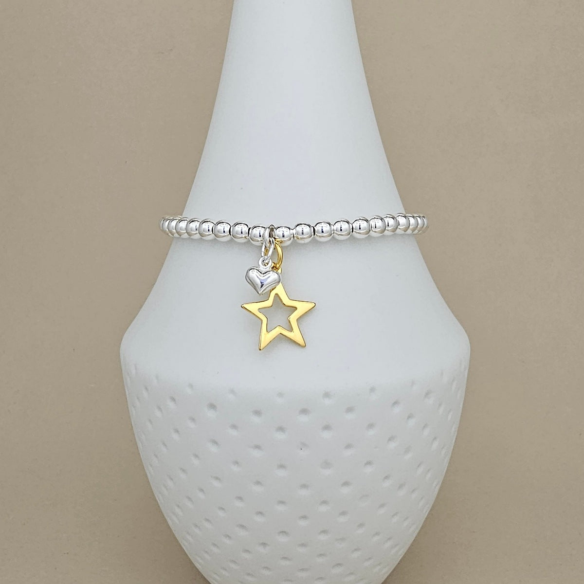 A silver beaded bracelet with a dangling gold star and silver heart charm, displayed on a white textured cone stand against a beige background.