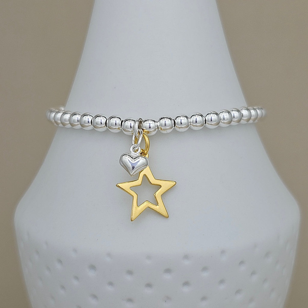 A silver beaded bracelet with a dangling charm featuring a polished silver heart and an openwork gold star, displayed on a white ceramic cone against a warm beige background.