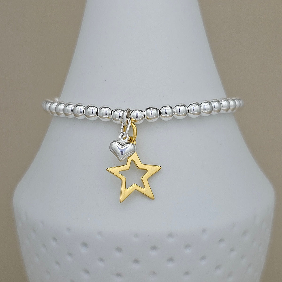 A silver beaded bracelet with a dangling charm featuring a polished silver heart and an openwork gold star, displayed on a white ceramic cone against a warm beige background.