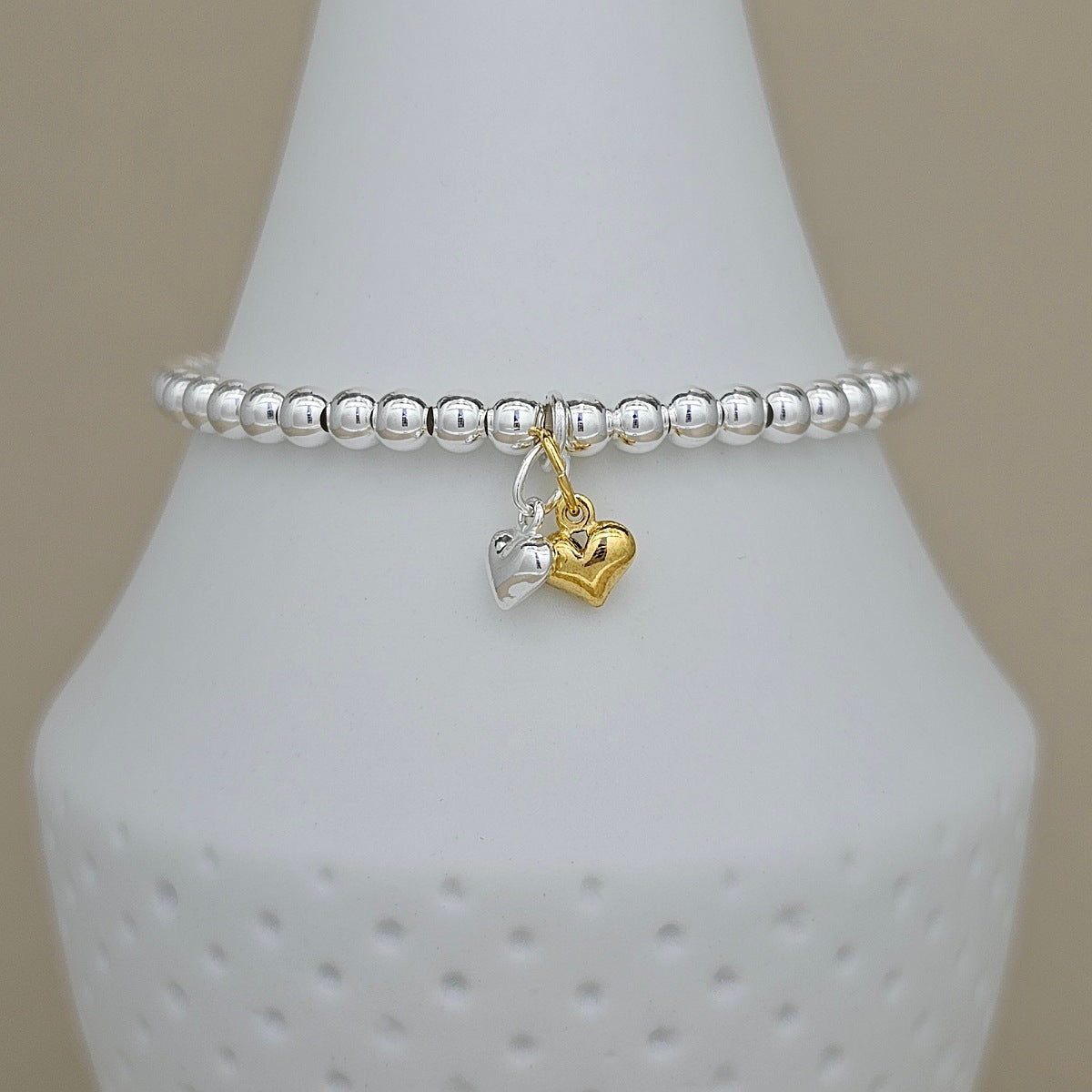 A silver beaded stretch bracelet with two dangling heart charms, one silver and one gold, displayed on a white cone-shaped jewelry stand against a beige background.