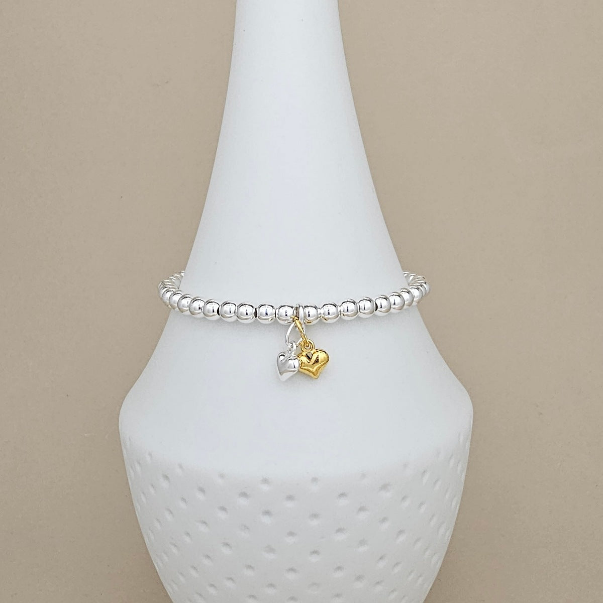 A silver beaded bracelet with two heart charms (one gold, one silver) displayed on a white cone jewelry stand against a beige background.