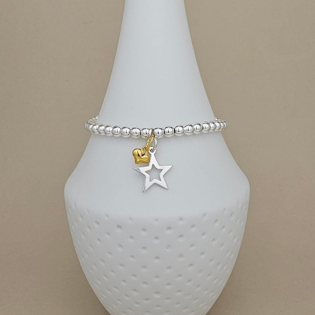 A silver beaded bracelet with a dangling star and heart charm, displayed on a white textured cone stand against a beige background.