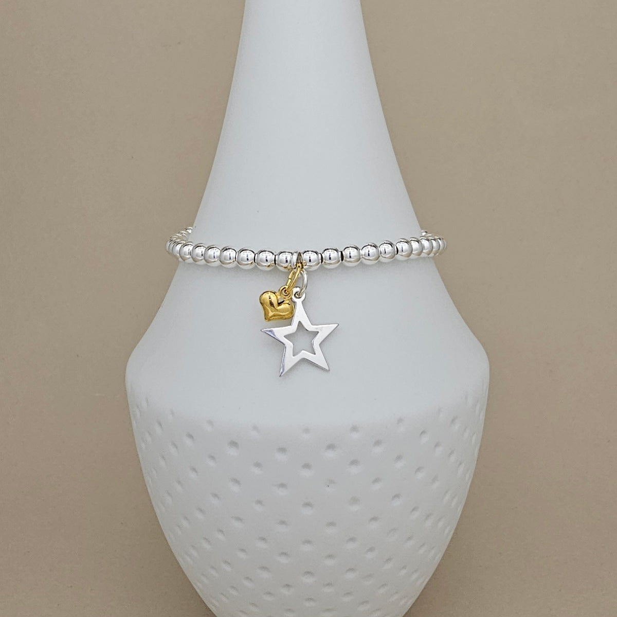 A silver beaded bracelet with a dangling star and heart charm, displayed on a white textured cone stand against a beige background.