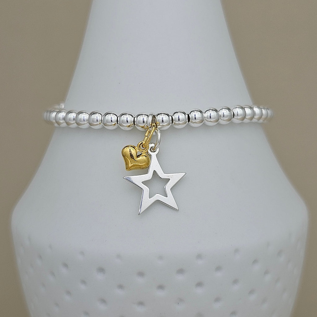 A silver beaded stretch bracelet with dangling charms, including a polished gold heart and a silver cutout star, displayed on a white ceramic jewelry stand.