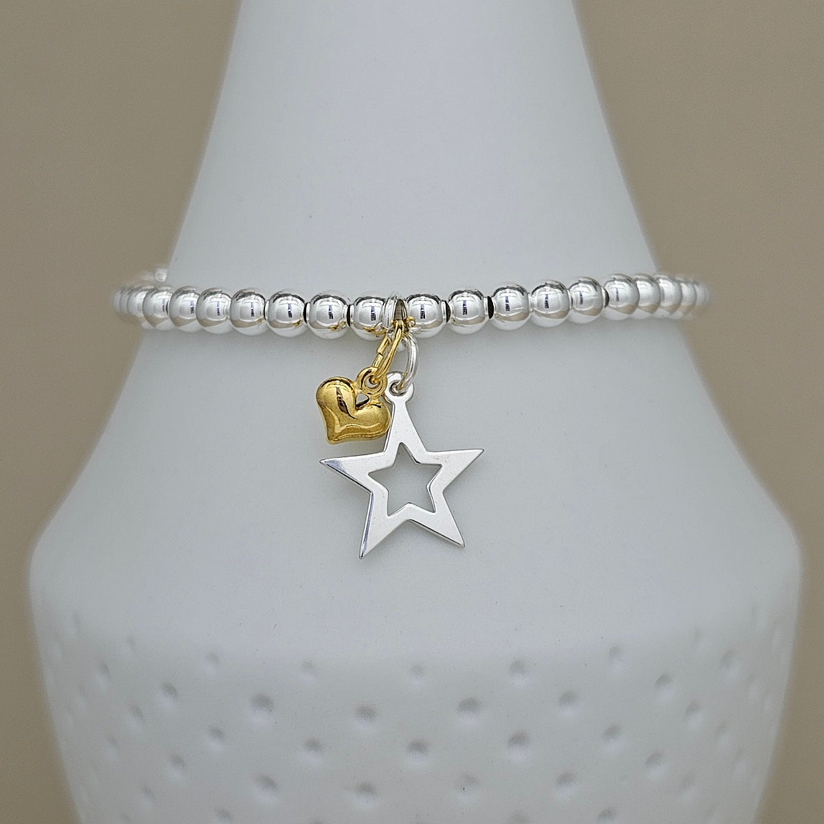 A silver beaded stretch bracelet with dangling charms, including a polished gold heart and a silver cutout star, displayed on a white ceramic jewelry stand.