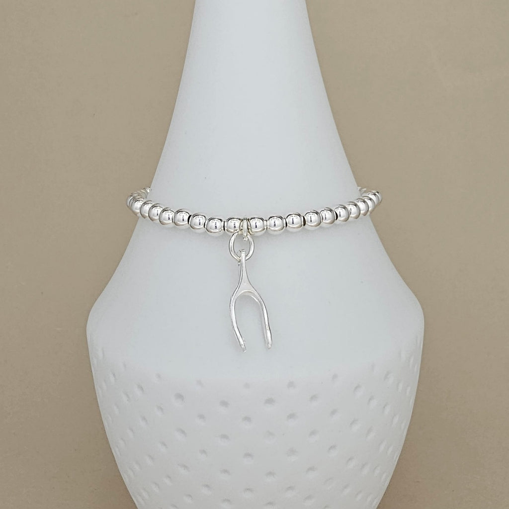 A silver beaded bracelet with a small hanging wishbone charm, displayed on a white ceramic cone stand against a warm beige background.