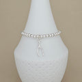 A silver beaded bracelet with a small hanging wishbone charm, displayed on a white ceramic cone stand against a warm beige background.