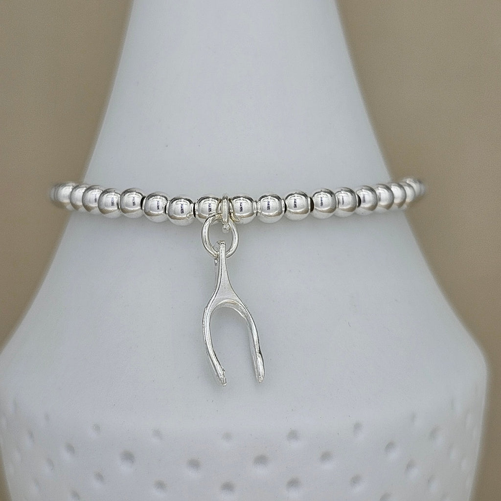 A silver beaded stretch bracelet with a small dangling wishbone charm, displayed on a white cone-shaped jewelry stand against a soft beige background.