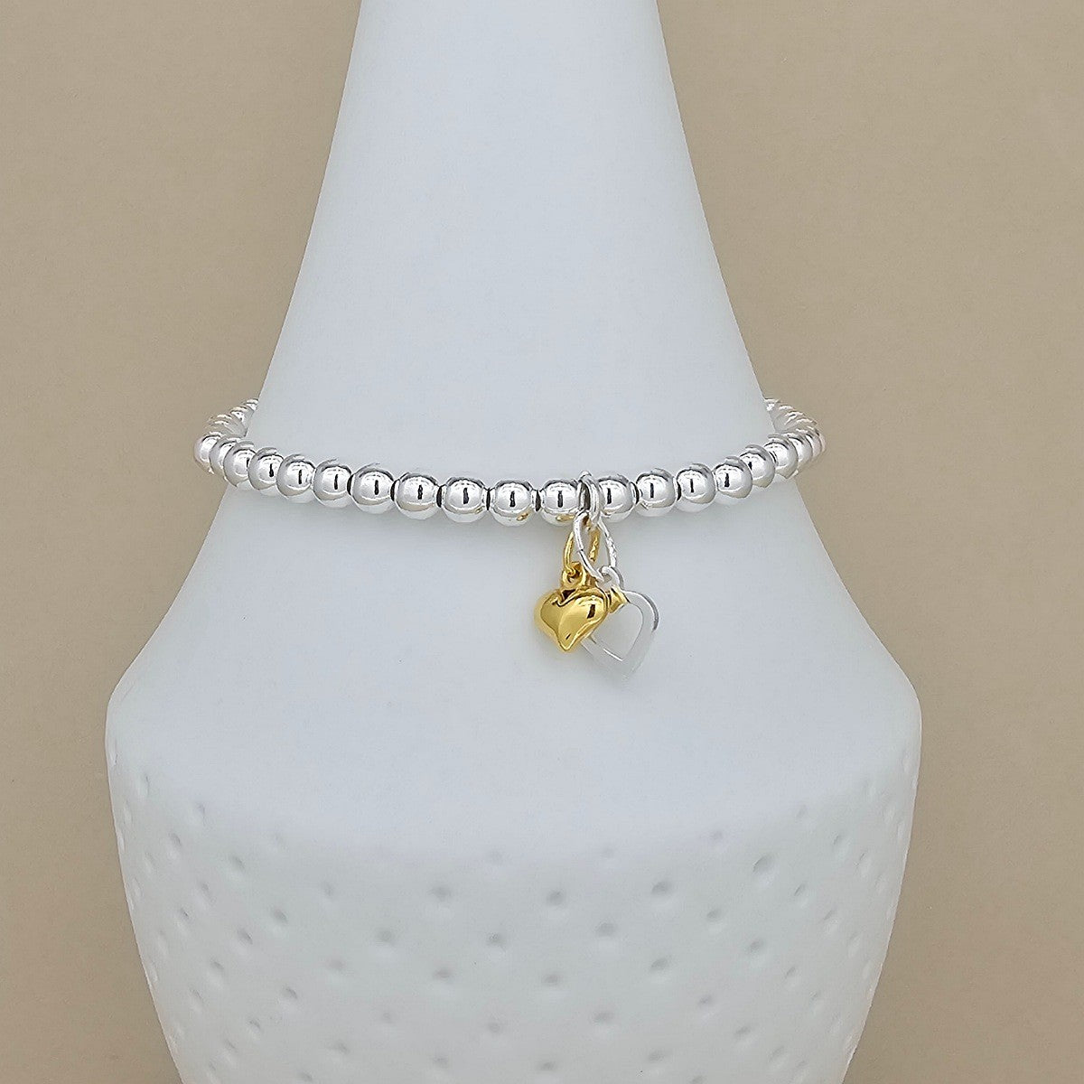 Silver beaded bracelet with a gold heart charm and silver open heart on a white mannequin arm against a beige background