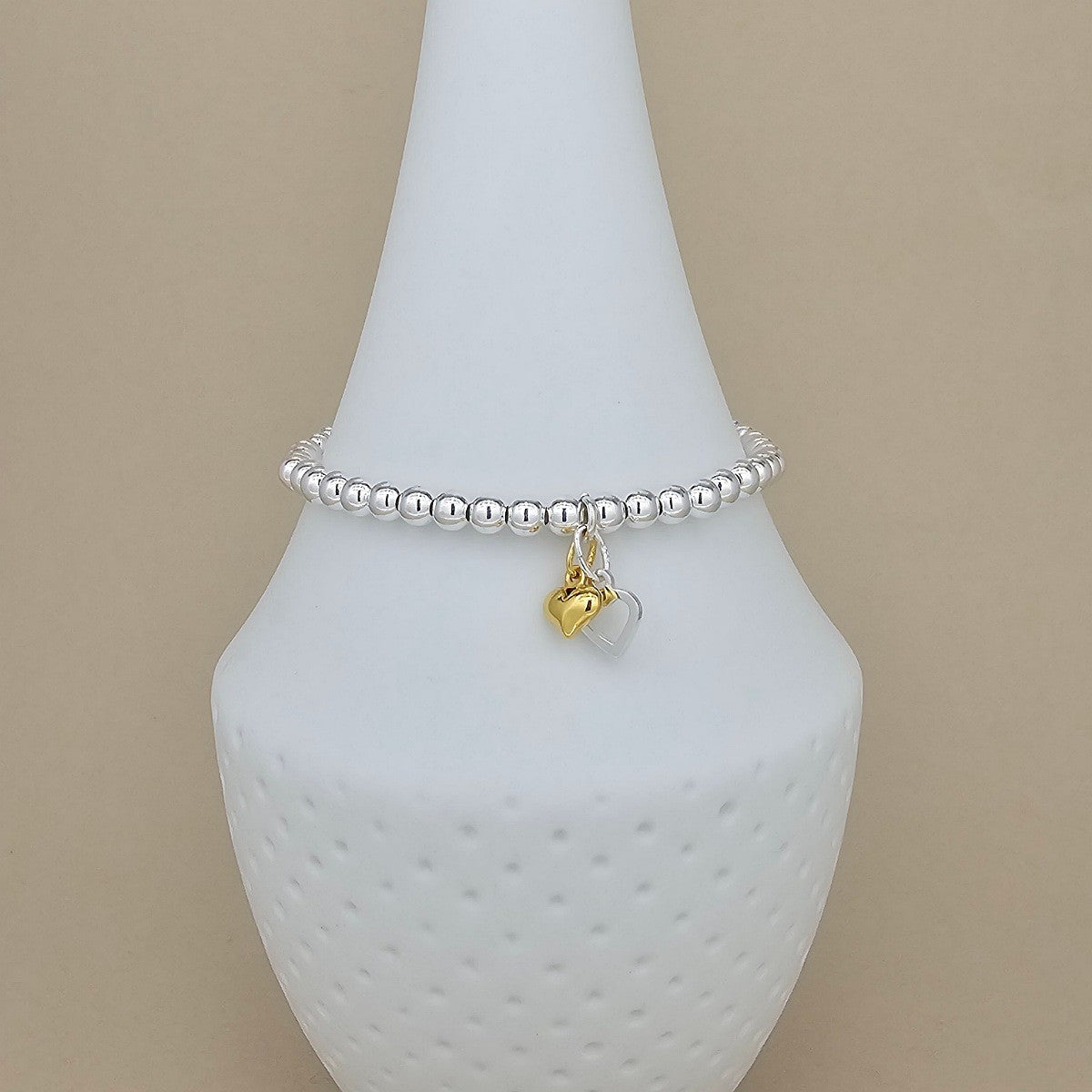A silver beaded bracelet with two dangling heart charms, one gold and one being a silver open heart, displayed on a white cone jewelry stand against a beige background.