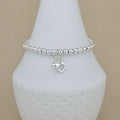 A silver beaded bracelet with a double heart charm, displayed on a white ceramic jewelry stand against a beige background.