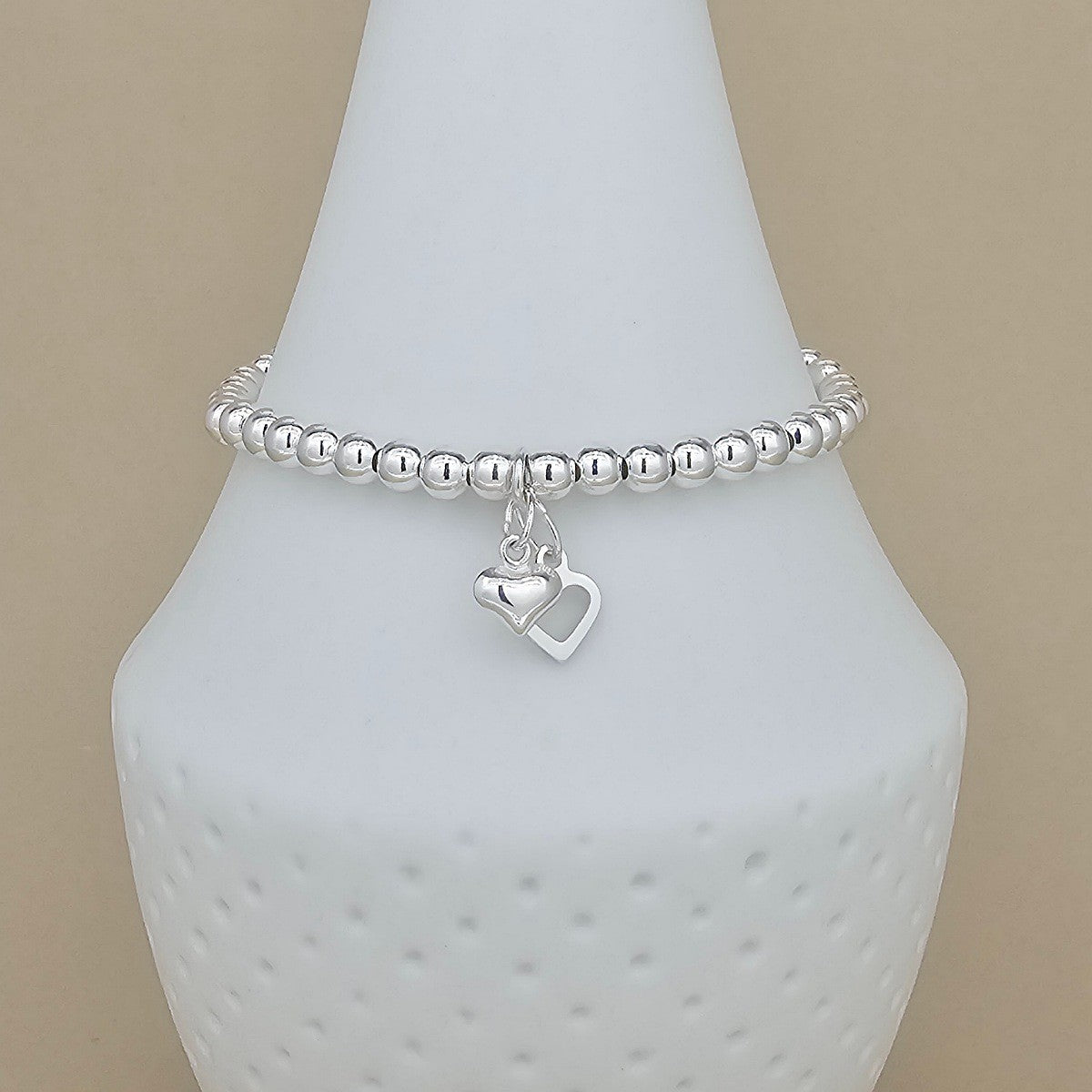 A silver beaded bracelet with a double heart charm, displayed on a white ceramic jewelry stand against a beige background.