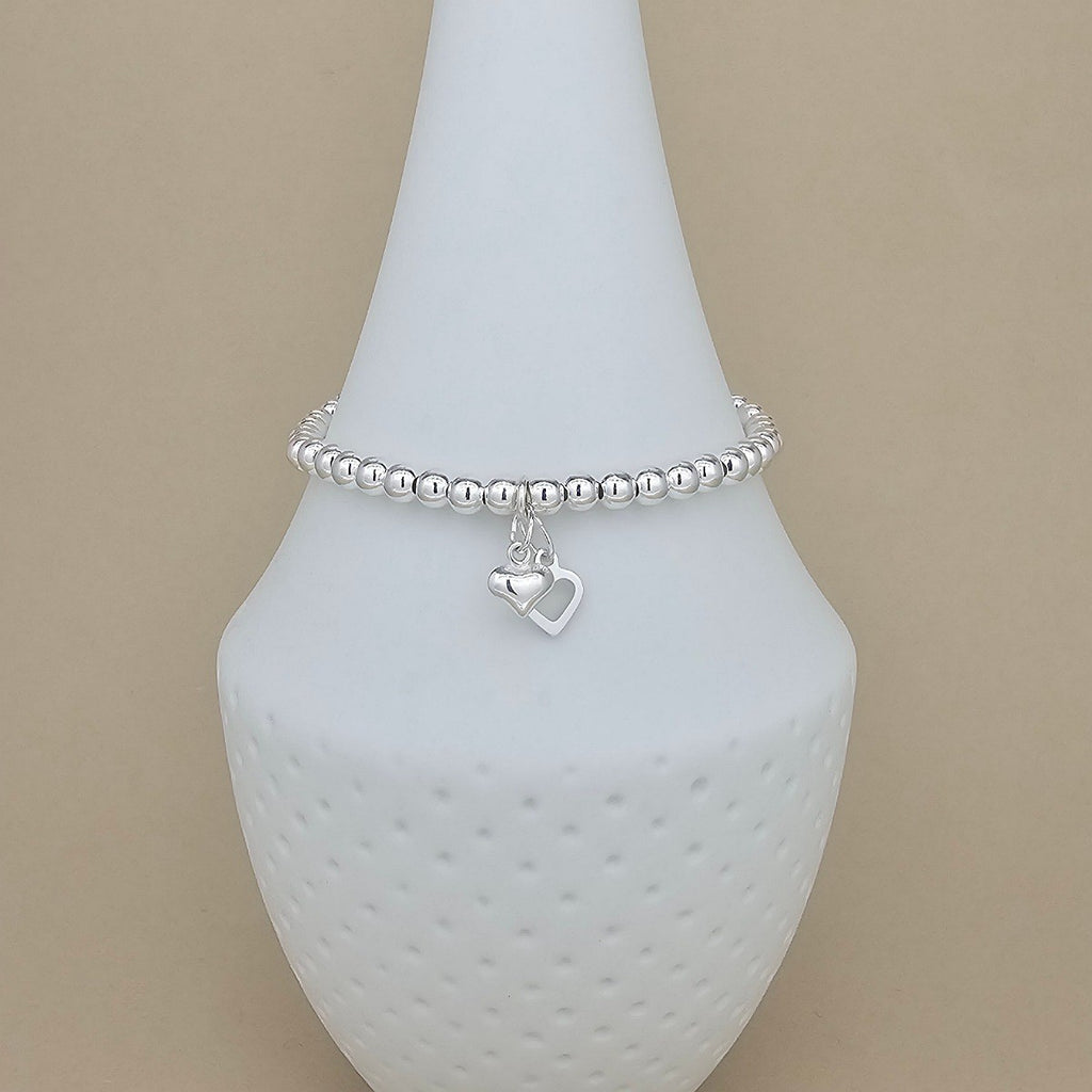A silver beaded bracelet with a dangling open heart charm, displayed on a white textured jewelry stand against a solid beige background.