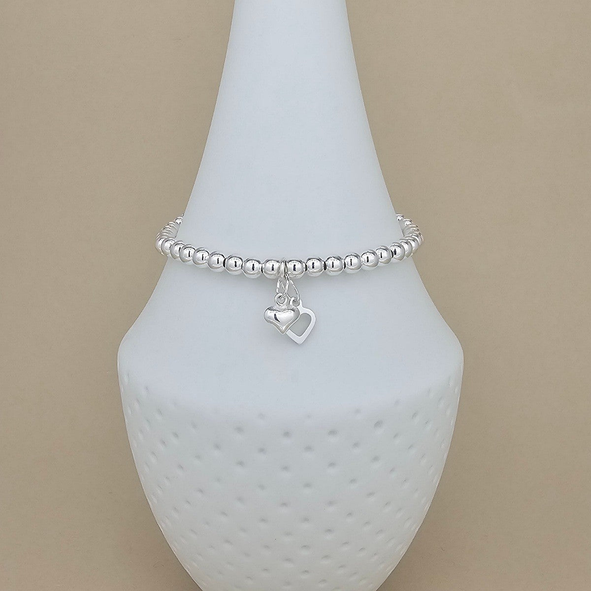 A silver beaded bracelet with a dangling open heart charm, displayed on a white textured jewelry stand against a solid beige background.