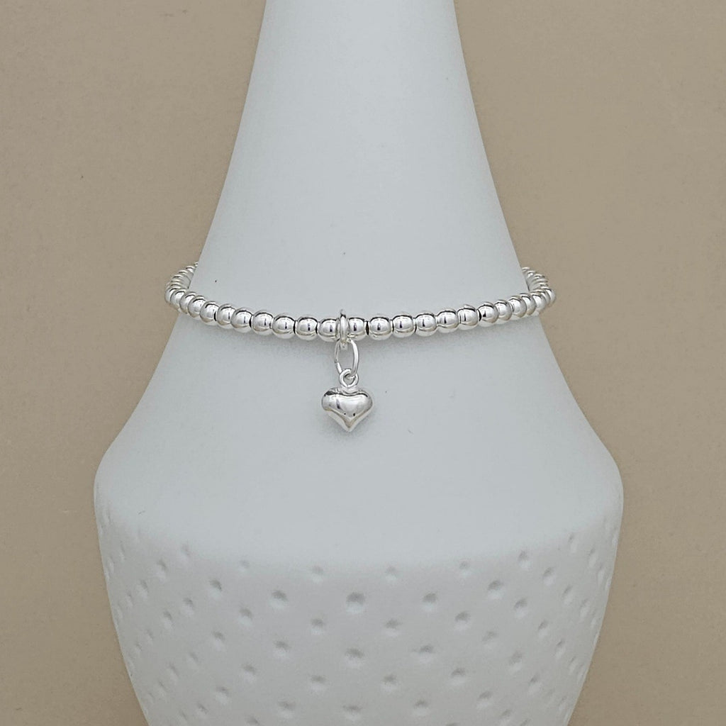 A silver beaded bracelet with a small heart charm, displayed on a white ceramic jewelry stand against a beige background.