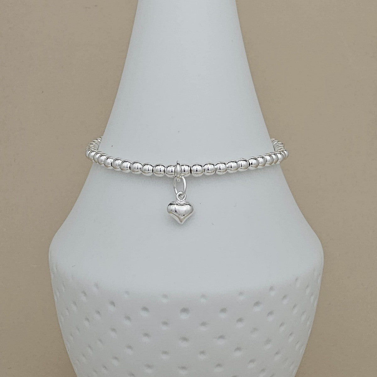 A silver beaded bracelet with a small heart charm, displayed on a white ceramic jewelry stand against a beige background.