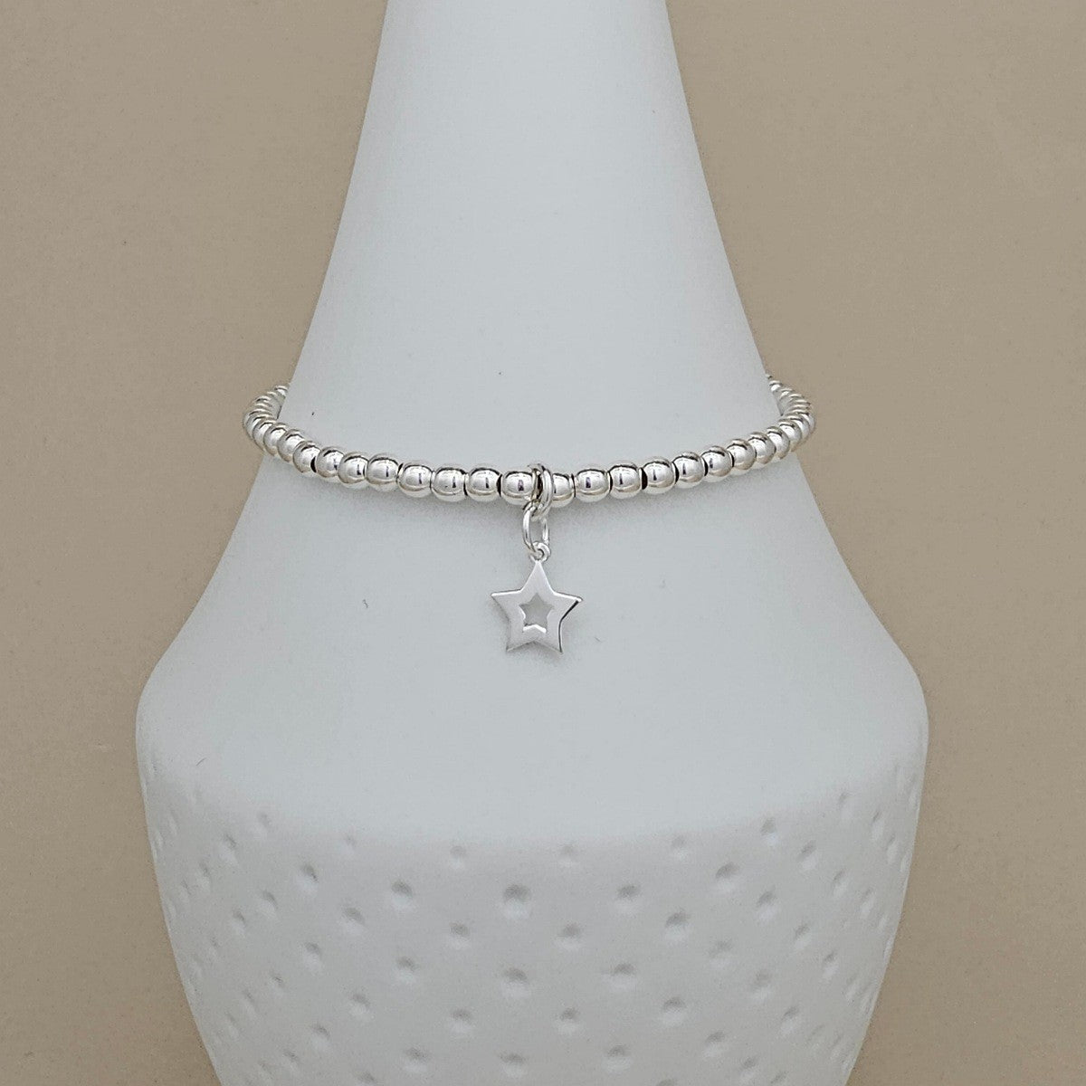A silver beaded bracelet with a dangling star charm, displayed on a white ceramic jewelry bust against a beige background.
