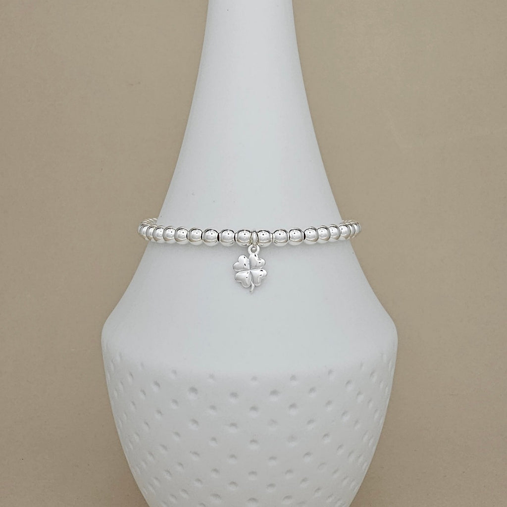 A silver beaded bracelet with a small dangling four-leaf clover charm, displayed on a white ceramic jewelry cone against a beige background.