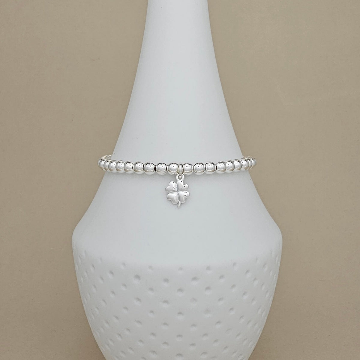 A silver beaded bracelet with a small dangling four-leaf clover charm, displayed on a white ceramic jewelry cone against a beige background.