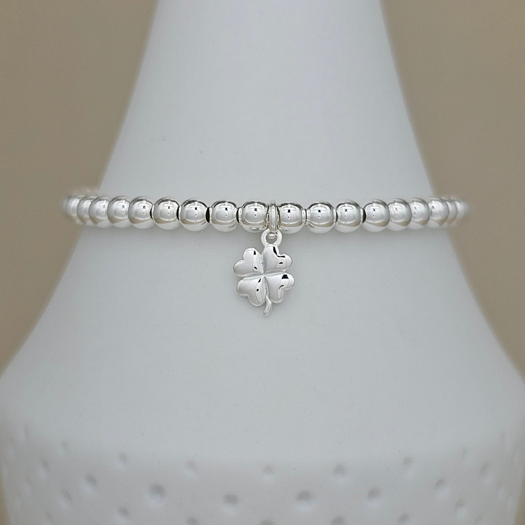 A silver beaded bracelet with a dangling four-leaf clover charm, displayed on a white ceramic jewelry cone against a neutral beige background.