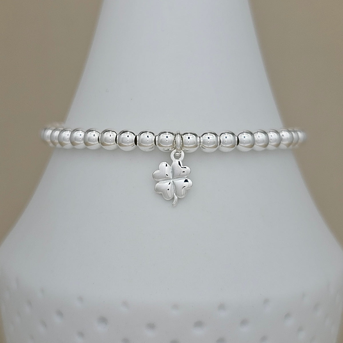 A silver beaded bracelet with a dangling four-leaf clover charm, displayed on a white ceramic jewelry cone against a neutral beige background.