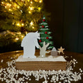 A handmade metal Christmas cutout scene featuring polar bear figure, rabbit and hedgehog, a decorated green Christmas tree, and a small star, displayed on a wooden base with white pebbles, with a blurred Christmas tree in the background